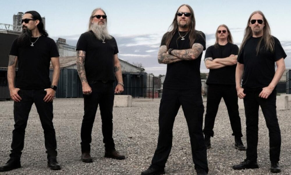 Five men in black shirts and sunglasses pose in an industrial yard with gravel ground and warehouses nearby.