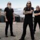 Five men in black shirts and sunglasses pose in an industrial yard with gravel ground and warehouses nearby.