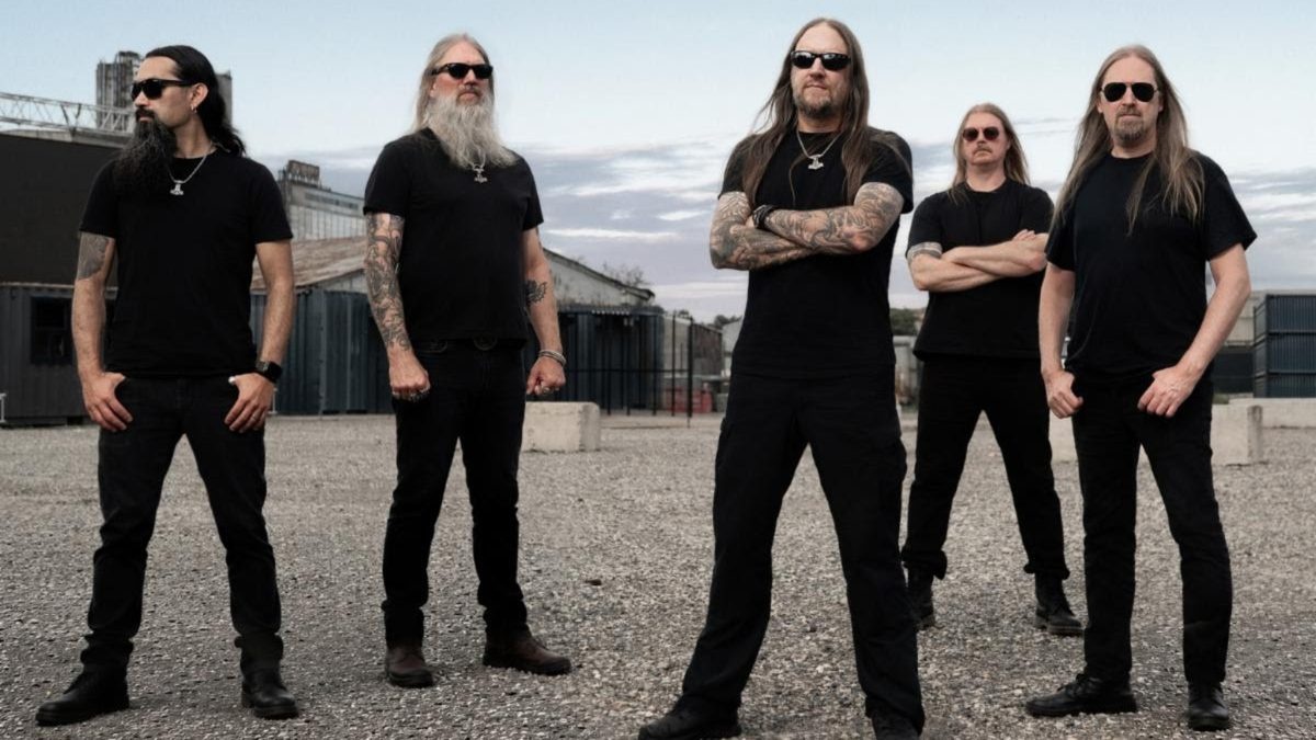 Five men in black shirts and sunglasses pose in an industrial yard with gravel ground and warehouses nearby.