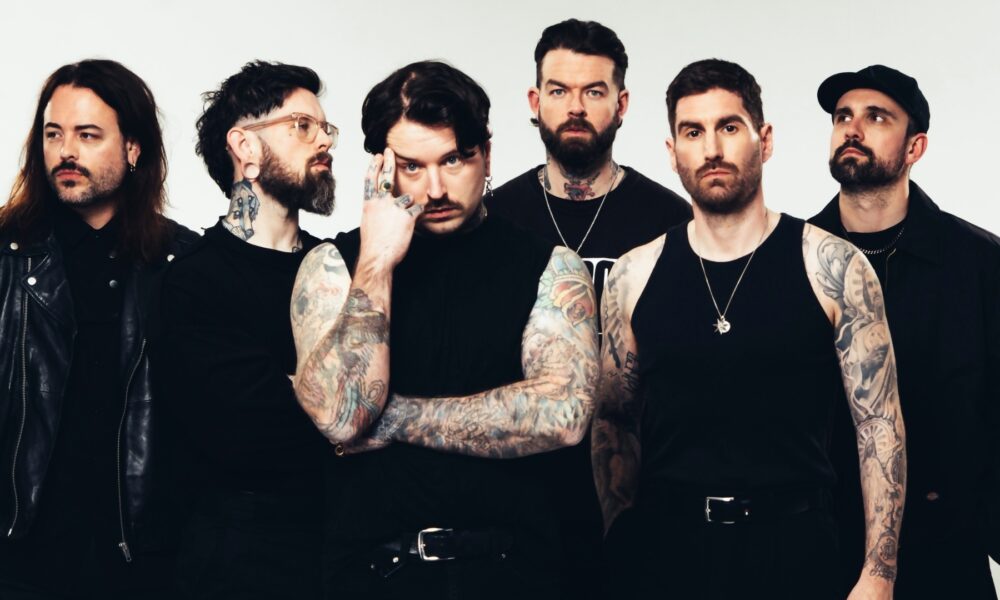 Six-member rock band posing in black outfits; heavily tattooed arms visible, standing against a light backdrop.