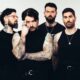 Six-member rock band posing in black outfits; heavily tattooed arms visible, standing against a light backdrop.