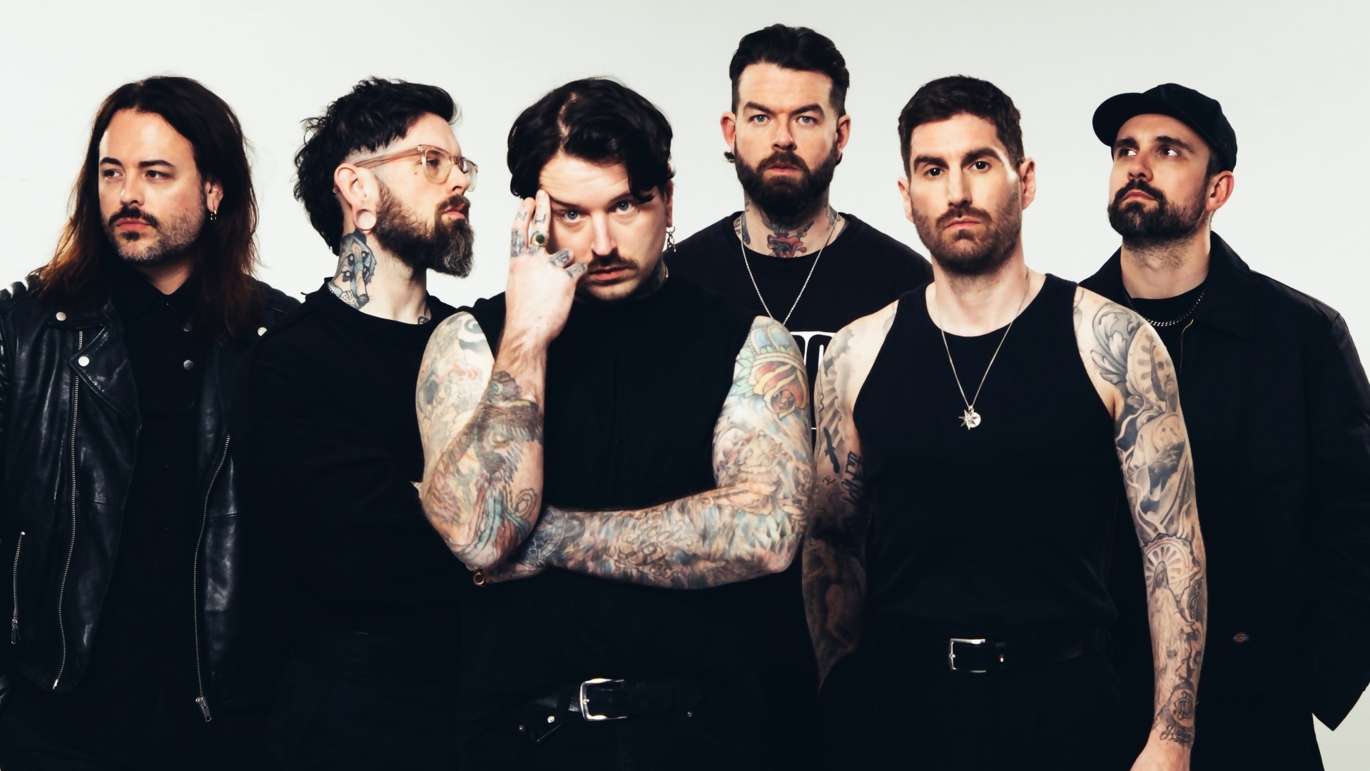 Six-member rock band posing in black outfits; heavily tattooed arms visible, standing against a light backdrop.