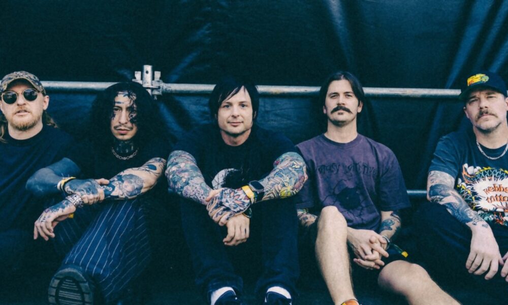 Five tattooed men sitting in a row against a dark backdrop, looking at the camera.
