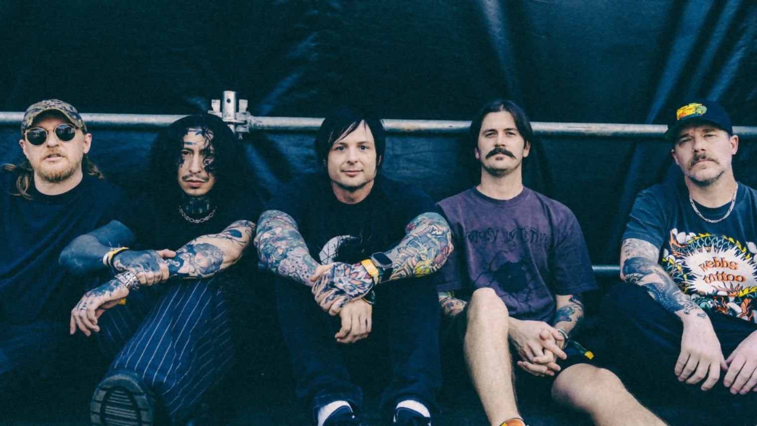 Five tattooed men sitting in a row against a dark backdrop, looking at the camera.