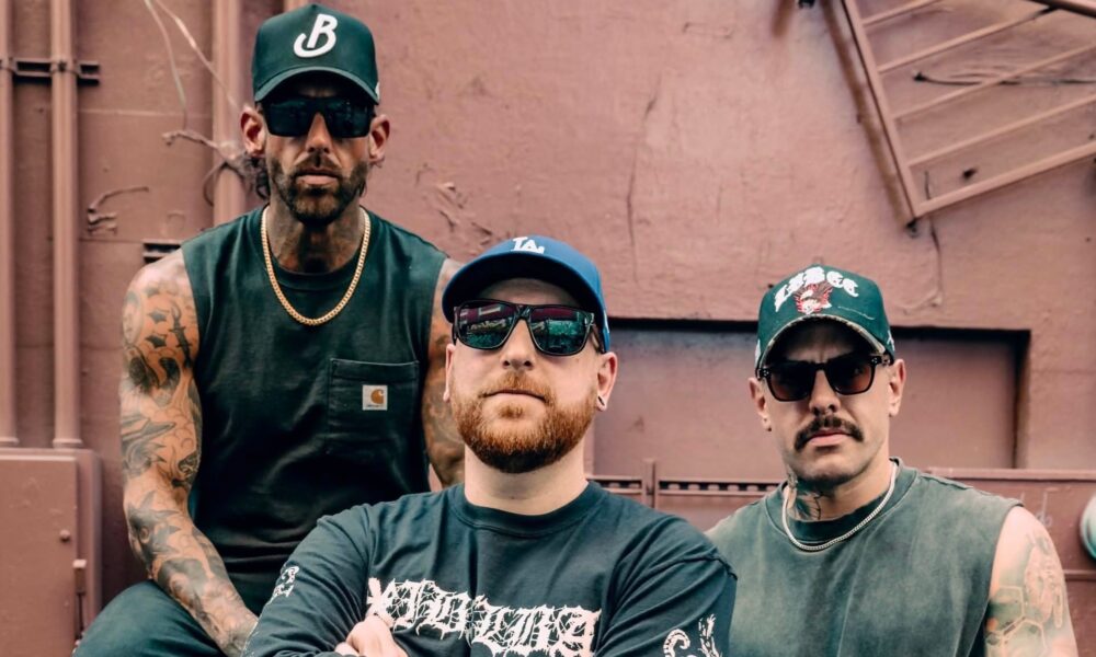 Three men in green shirts and sunglasses pose together outdoors in front of a brown industrial wall.