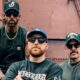 Three men in green shirts and sunglasses pose together outdoors in front of a brown industrial wall.