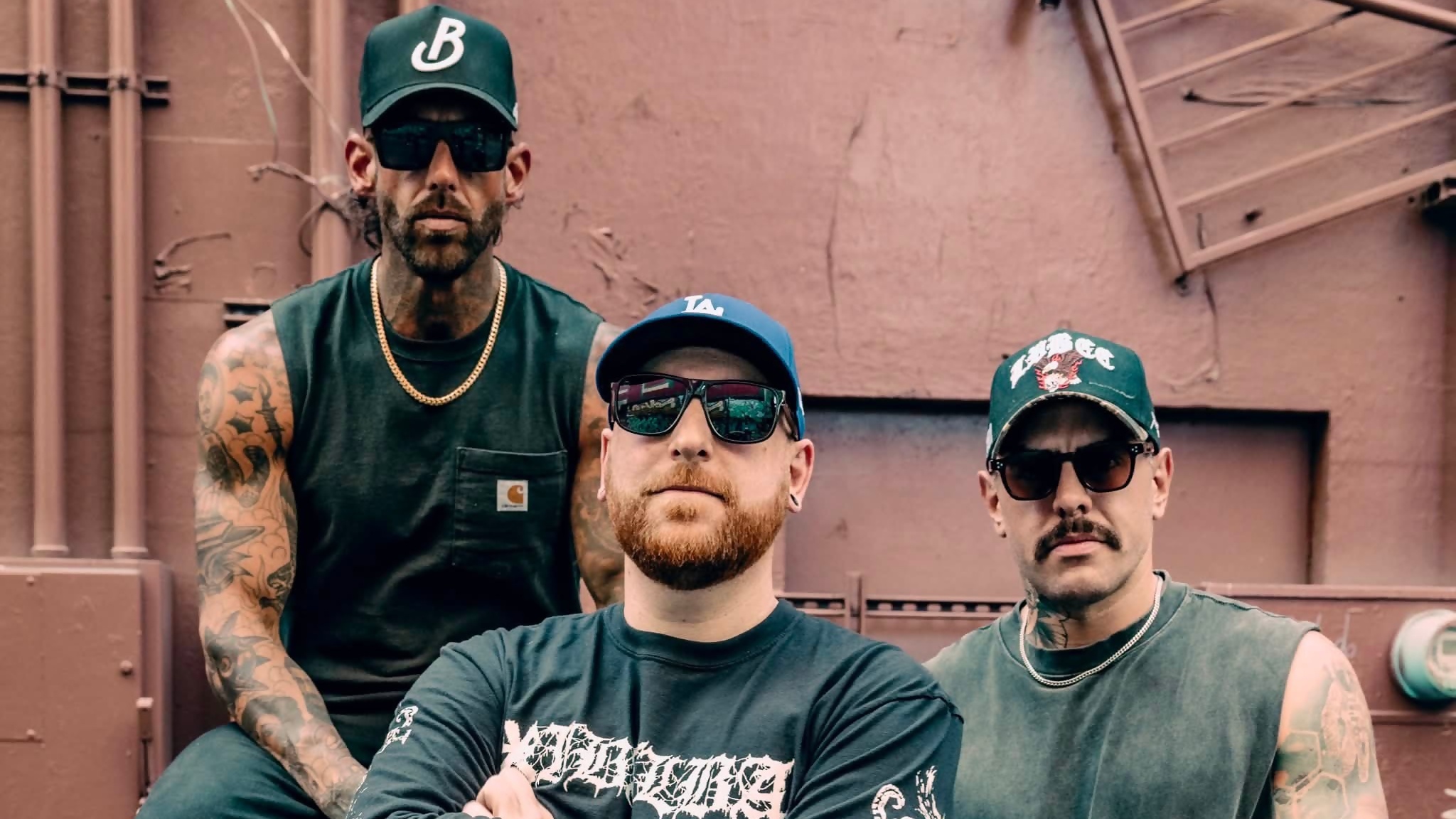 Three men in green shirts and sunglasses pose together outdoors in front of a brown industrial wall.