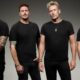 Four men in black T-shirts stand side by side, facing the camera against a gray studio backdrop.