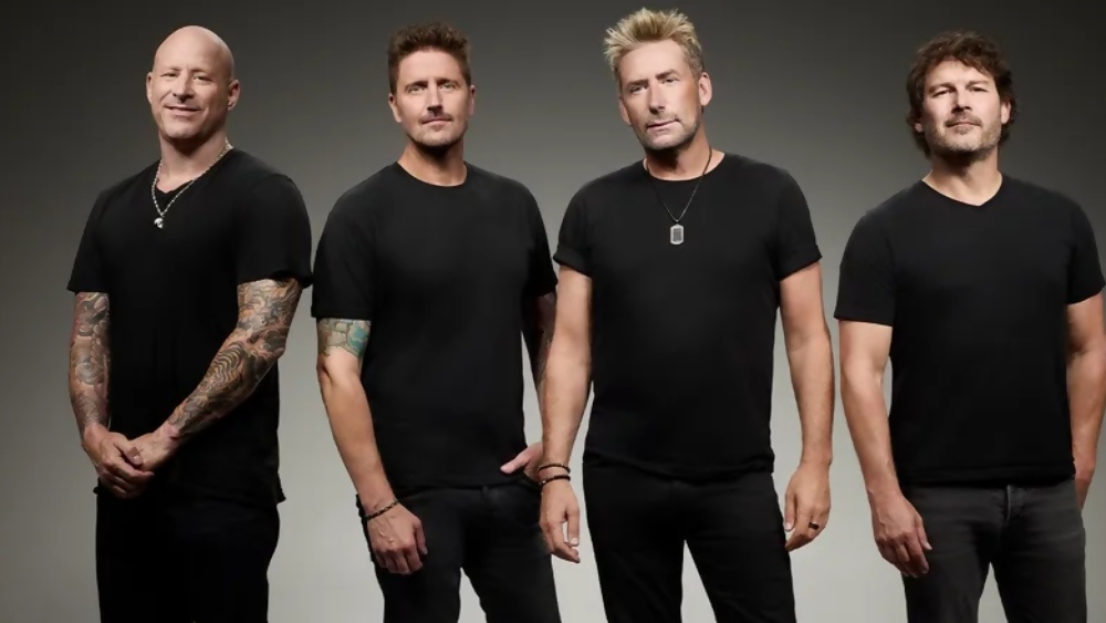 Four men in black T-shirts stand side by side, facing the camera against a gray studio backdrop.